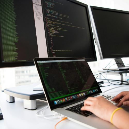 A developer writes code on a laptop in front of multiple monitors in an office setting.